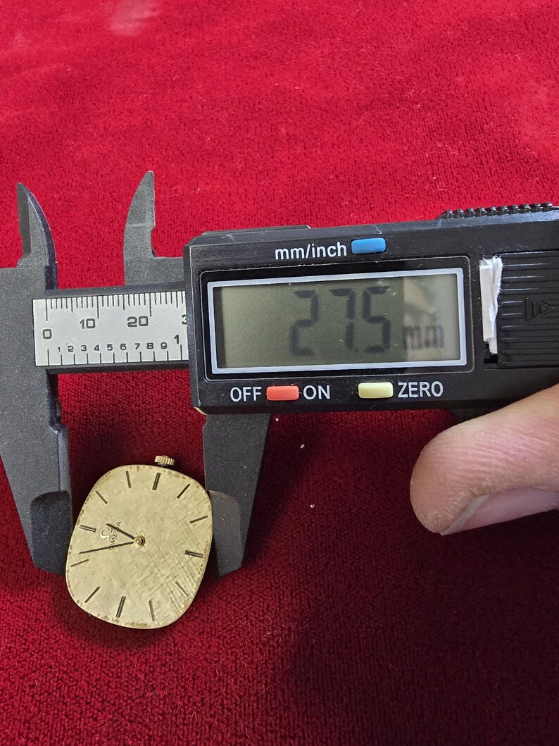 May include: A gold-toned watch face is measured with a digital caliper. The caliper displays 27.5 mm. The watch face has a textured surface, black hands, and black hour markers. The watch face is oval-shaped and sits on a red surface.