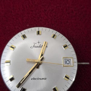 Puede incluir: Primer plano de una esfera de reloj plateada con manecillas y marcadores dorados. La esfera del reloj tiene la palabra "Tradition" en cursiva y "electronic" impresa debajo. La fecha se muestra como "10" en una peque&ntilde;a ventana. "Swiss Made" est&aacute; impreso en la parte inferior.