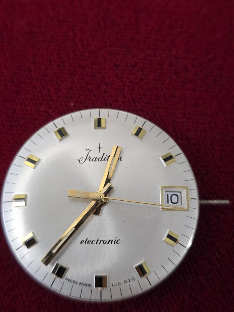 Puede incluir: Primer plano de una esfera de reloj plateada con manecillas y marcadores dorados. La esfera del reloj tiene la palabra "Tradition" en cursiva y "electronic" impresa debajo. La fecha se muestra como "10" en una peque&ntilde;a ventana. "Swiss Made" est&aacute; impreso en la parte inferior.