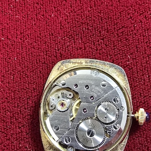 May include: Close-up of a vintage gold-toned watch with the back removed, revealing the intricate mechanical movement. The watch face is rectangular with a gold bezel. The internal gears and components are visible, with the brand name "EBEL" and "107" etched on a circular piece.