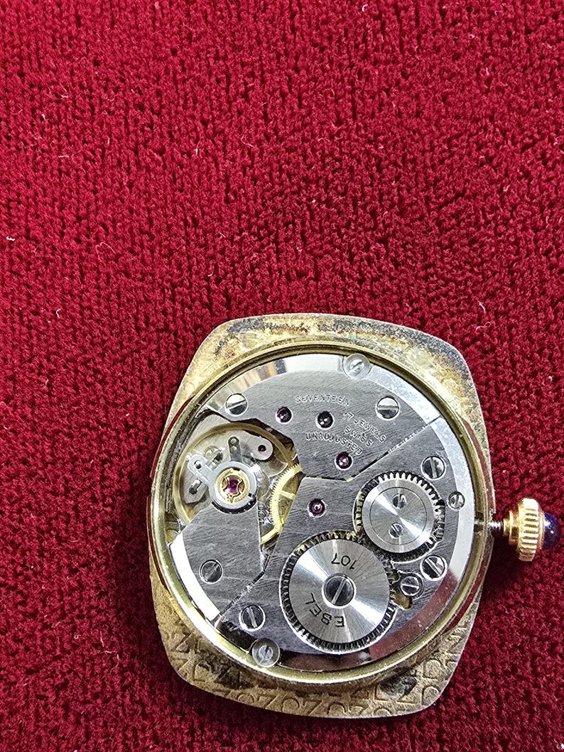 May include: Close-up of a vintage gold-toned watch with the back removed, revealing the intricate mechanical movement. The watch face is rectangular with a gold bezel. The internal gears and components are visible, with the brand name "EBEL" and "107" etched on a circular piece.