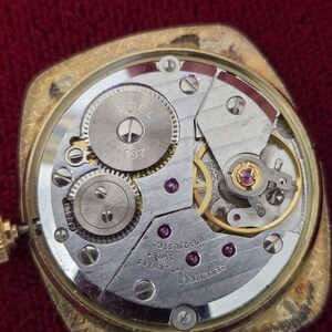 May include: Close-up of a vintage gold-toned watch mechanism. The watch's inner workings are visible, including gears, jewels, and the "EBEL" logo. The watch is labeled "17 Jewels Swiss" and "Unadjusted".