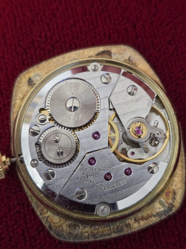 May include: Close-up of a vintage gold-toned watch mechanism. The watch's inner workings are visible, including gears, jewels, and the "EBEL" logo. The watch is labeled "17 Jewels Swiss" and "Unadjusted".
