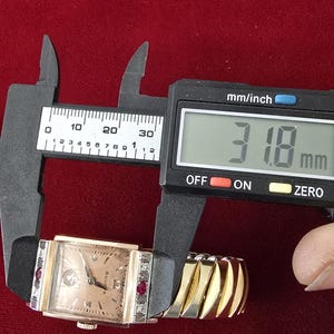 May include: A vintage Bulova watch with a rectangular face, featuring a light-colored dial and diamond and ruby accents. The watch is measured with a digital caliper, showing a width of 31.8 mm. The watch has a gold-toned expanding band and a black leather strap.