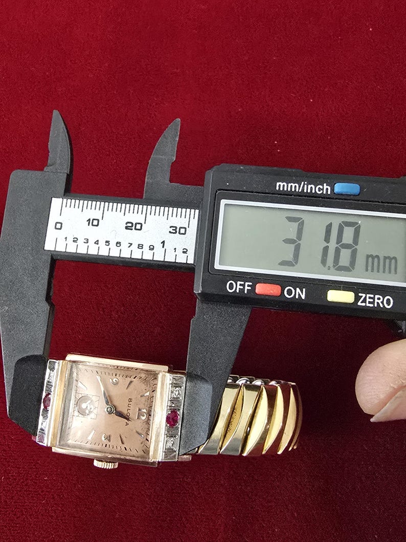 May include: A vintage Bulova watch with a rectangular face, featuring a light-colored dial and diamond and ruby accents. The watch is measured with a digital caliper, showing a width of 31.8 mm. The watch has a gold-toned expanding band and a black leather strap.
