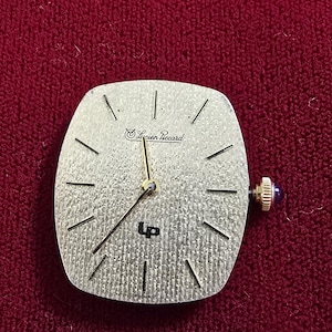 May include: A silver-toned watch face with the text "Lucien Piccard" and "UP" is displayed. The watch face has black lines indicating the hours and gold-colored hands. A small, blue-jeweled knob is on the side. The watch face is square with rounded edges.