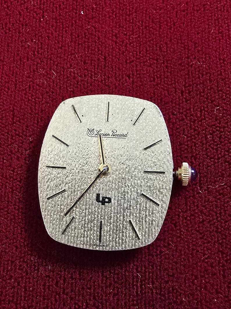 May include: A silver-toned watch face with the text "Lucien Piccard" and "UP" is displayed. The watch face has black lines indicating the hours and gold-colored hands. A small, blue-jeweled knob is on the side. The watch face is square with rounded edges.
