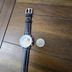 Op de afbeelding: Een polshorloge met een zwarte leren band met witte contraststiksels en een zilverkleurige kast. De wijzerplaat toont een gele zon en een paarse maan. Een zilverkleurige munt ligt naast het horloge voor de schaalvergroting.