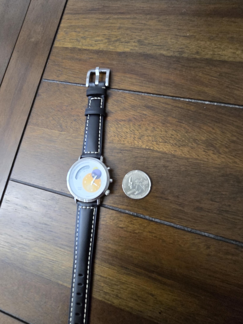 Op de afbeelding: Een polshorloge met een zwarte leren band met witte contraststiksels en een zilverkleurige kast. De wijzerplaat toont een gele zon en een paarse maan. Een zilverkleurige munt ligt naast het horloge voor de schaalvergroting.