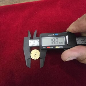 K&ouml;nnte beinhalten: Ein kleines, rundes Zifferblatt mit cremefarbenem Zifferblatt und goldenen Zeigern und Markierungen. Ein digitaler Messschieber misst das Zifferblatt mit 18,8 mm. Der Hintergrund ist ein tiefroter Stoff. Das Zifferblatt hat r&ouml;mische Ziffern.