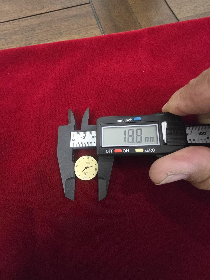 K&ouml;nnte beinhalten: Ein kleines, rundes Zifferblatt mit cremefarbenem Zifferblatt und goldenen Zeigern und Markierungen. Ein digitaler Messschieber misst das Zifferblatt mit 18,8 mm. Der Hintergrund ist ein tiefroter Stoff. Das Zifferblatt hat r&ouml;mische Ziffern.