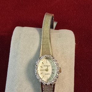 K&ouml;nnte beinhalten: Eine goldfarbene Armitron-Quarzuhr mit wei&szlig;em Zifferblatt und einer mit kleinen, klaren Steinen besetzten L&uuml;nette. Die Uhr hat ein Armband im Mesh-Stil und wird auf hellem Untergrund vor rotem Hintergrund pr&auml;sentiert.