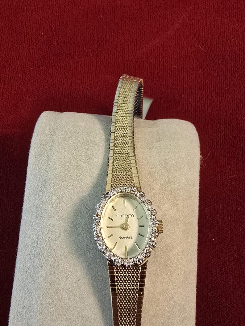 K&ouml;nnte beinhalten: Eine goldfarbene Armitron-Quarzuhr mit wei&szlig;em Zifferblatt und einer mit kleinen, klaren Steinen besetzten L&uuml;nette. Die Uhr hat ein Armband im Mesh-Stil und wird auf hellem Untergrund vor rotem Hintergrund pr&auml;sentiert.