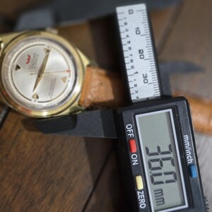 May include: A gold-toned wristwatch with a light-colored face and a brown leather band. The watch is being measured with a digital caliper, showing a measurement of 36.0 mm.