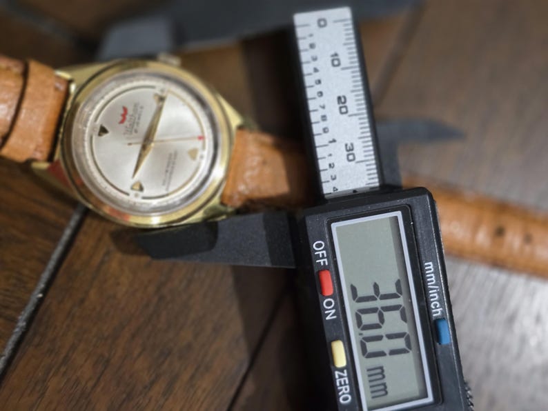 May include: A gold-toned wristwatch with a light-colored face and a brown leather band. The watch is being measured with a digital caliper, showing a measurement of 36.0 mm.