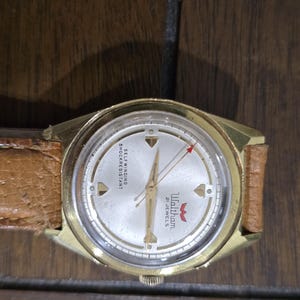 May include: A vintage gold-toned Waltham wristwatch with a light-colored face and a brown leather band. The watch face features the text "Waltham 21 JEWELS" and "SELF WINDING SHOCKRESISTANT". The second hand has a red arrow.