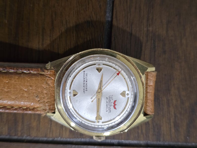 May include: A vintage gold-toned Waltham wristwatch with a light-colored face and a brown leather band. The watch face features the text "Waltham 21 JEWELS" and "SELF WINDING SHOCKRESISTANT". The second hand has a red arrow.