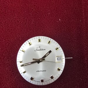 Puede incluir: Una esfera de reloj plateada con la palabra "Tradition" encima del centro. La esfera del reloj tiene manecillas negras y marcadores dorados. La palabra "electronic" est&aacute; impresa debajo. La fecha se muestra como "10".