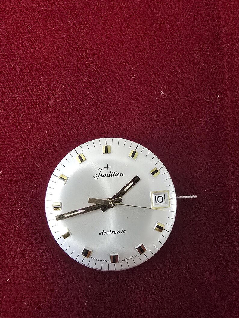 Puede incluir: Una esfera de reloj plateada con la palabra "Tradition" encima del centro. La esfera del reloj tiene manecillas negras y marcadores dorados. La palabra "electronic" est&aacute; impresa debajo. La fecha se muestra como "10".