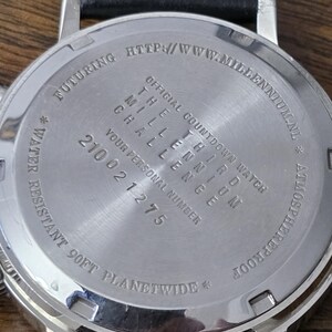 Op de afbeelding: De roestvrijstalen achterkant van een horlogekast met een zwarte leren band. De gegraveerde tekst luidt: OFFICIAL COUNTDOWN WATCH THE THIRD MILLENNIUM CHALLENGE, YOUR PERSONAL NUMBER 210021275, WATER RESISTANT 90FT PLANETWIDE en HTTP://WWW.MILLENNIUM.NL.