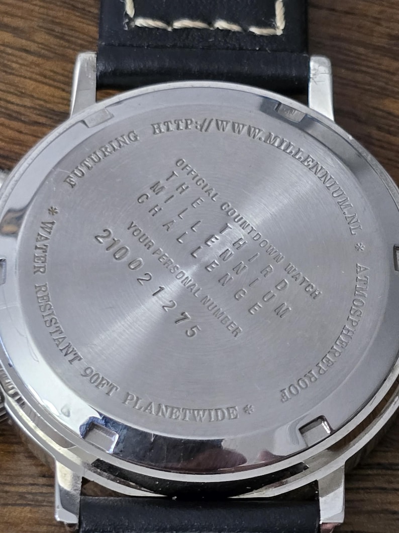 Op de afbeelding: De roestvrijstalen achterkant van een horlogekast met een zwarte leren band. De gegraveerde tekst luidt: OFFICIAL COUNTDOWN WATCH THE THIRD MILLENNIUM CHALLENGE, YOUR PERSONAL NUMBER 210021275, WATER RESISTANT 90FT PLANETWIDE en HTTP://WWW.MILLENNIUM.NL.