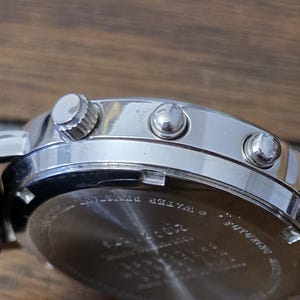 Op de afbeelding: Een zilverkleurige roestvrijstalen horlogekast met een zijdelingse kroon en twee drukknoppen. De achterkant van de horlogekast is van metaal en bevat gegraveerde tekst, waaronder de woorden WATER RESISTANT en andere gedeeltelijk zichtbare tekens.