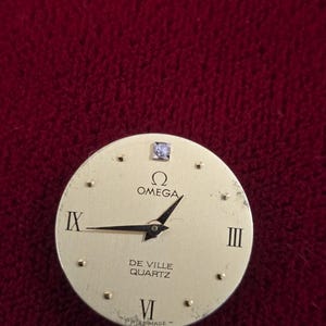 K&ouml;nnte beinhalten: Ein goldfarbenes Omega De Ville Quartz Zifferblatt mit schwarzen Zeigern und r&ouml;mischen Ziffern. Das Zifferblatt hat einen kleinen Diamanten bei 12 Uhr. Die W&ouml;rter "Omega", "De Ville", "Quartz" und "Swiss Made" sind aufgedruckt.