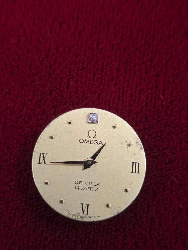 K&ouml;nnte beinhalten: Ein goldfarbenes Omega De Ville Quartz Zifferblatt mit schwarzen Zeigern und r&ouml;mischen Ziffern. Das Zifferblatt hat einen kleinen Diamanten bei 12 Uhr. Die W&ouml;rter "Omega", "De Ville", "Quartz" und "Swiss Made" sind aufgedruckt.