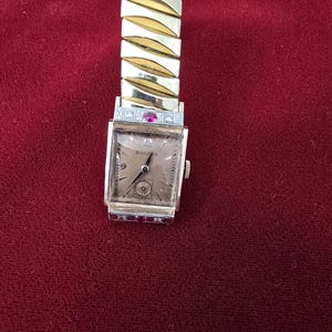May include: A vintage Bulova wristwatch with a gold-toned rectangular face and a gold-toned expanding band. The watch face has a light-colored background with black numerals and hands. Small red stones are set on the watch face.