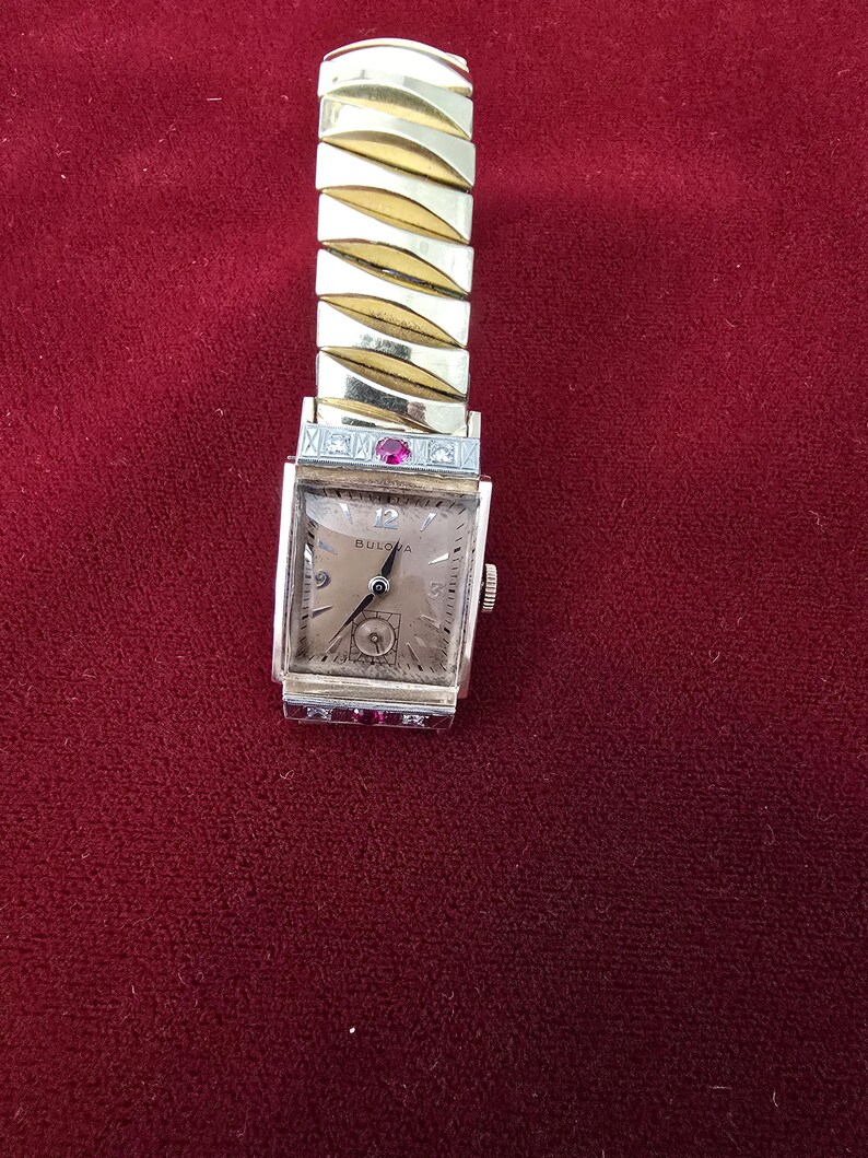 May include: A vintage Bulova wristwatch with a gold-toned rectangular face and a gold-toned expanding band. The watch face has a light-colored background with black numerals and hands. Small red stones are set on the watch face.