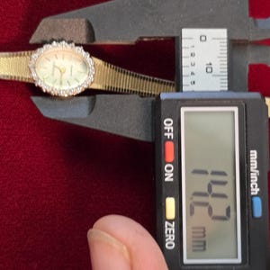 K&ouml;nnte beinhalten: Eine goldfarbene Armbanduhr mit ovalem Zifferblatt und einer L&uuml;nette, die mit kleinen, klaren Steinen besetzt ist. Das Zifferblatt ist hell mit schwarzen Zeigern und Markierungen. Das Armband ist aus goldfarbenem Gewebe. Das Messschieber zeigt 1,42 cm.