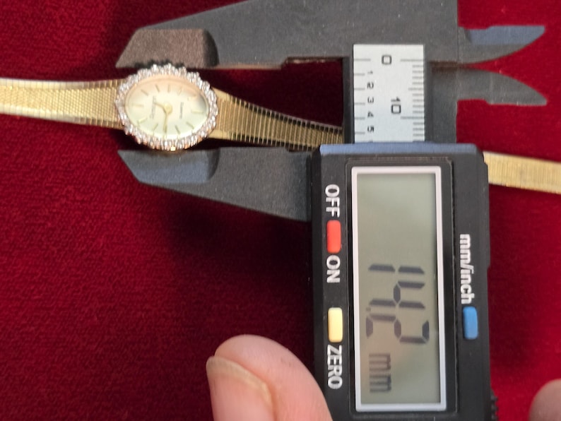 K&ouml;nnte beinhalten: Eine goldfarbene Armbanduhr mit ovalem Zifferblatt und einer L&uuml;nette, die mit kleinen, klaren Steinen besetzt ist. Das Zifferblatt ist hell mit schwarzen Zeigern und Markierungen. Das Armband ist aus goldfarbenem Gewebe. Das Messschieber zeigt 1,42 cm.