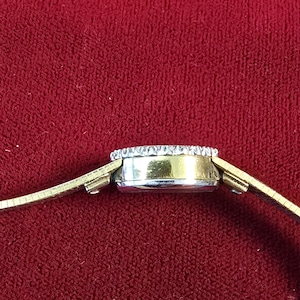K&ouml;nnte beinhalten: Seitenansicht einer goldfarbenen Armbanduhr mit rundem Zifferblatt. Das Zifferblatt ist mit kleinen, klaren Edelsteinen umrandet. Das Armband ist aus d&uuml;nnem, flexiblem Goldmetall. Der Hintergrund ist ein tiefes, einfarbiges Rot.