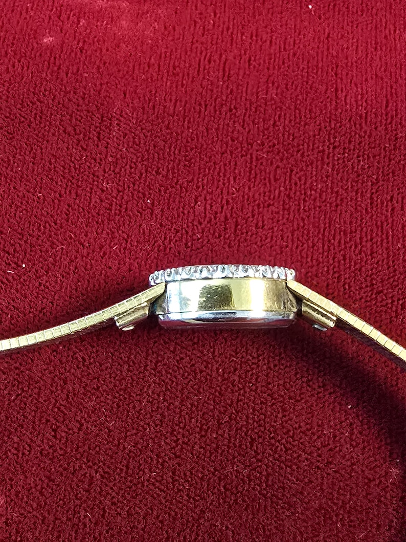 K&ouml;nnte beinhalten: Seitenansicht einer goldfarbenen Armbanduhr mit rundem Zifferblatt. Das Zifferblatt ist mit kleinen, klaren Edelsteinen umrandet. Das Armband ist aus d&uuml;nnem, flexiblem Goldmetall. Der Hintergrund ist ein tiefes, einfarbiges Rot.