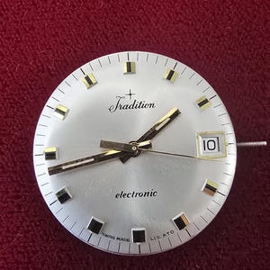 Puede incluir: Esfera de reloj plateada con las palabras "Tradition" y "electronic" impresas. La esfera del reloj tiene manecillas y marcadores dorados, con una ventana de fecha que muestra el n&uacute;mero "10". Las palabras "SWISS MADE" tambi&eacute;n est&aacute;n impresas.