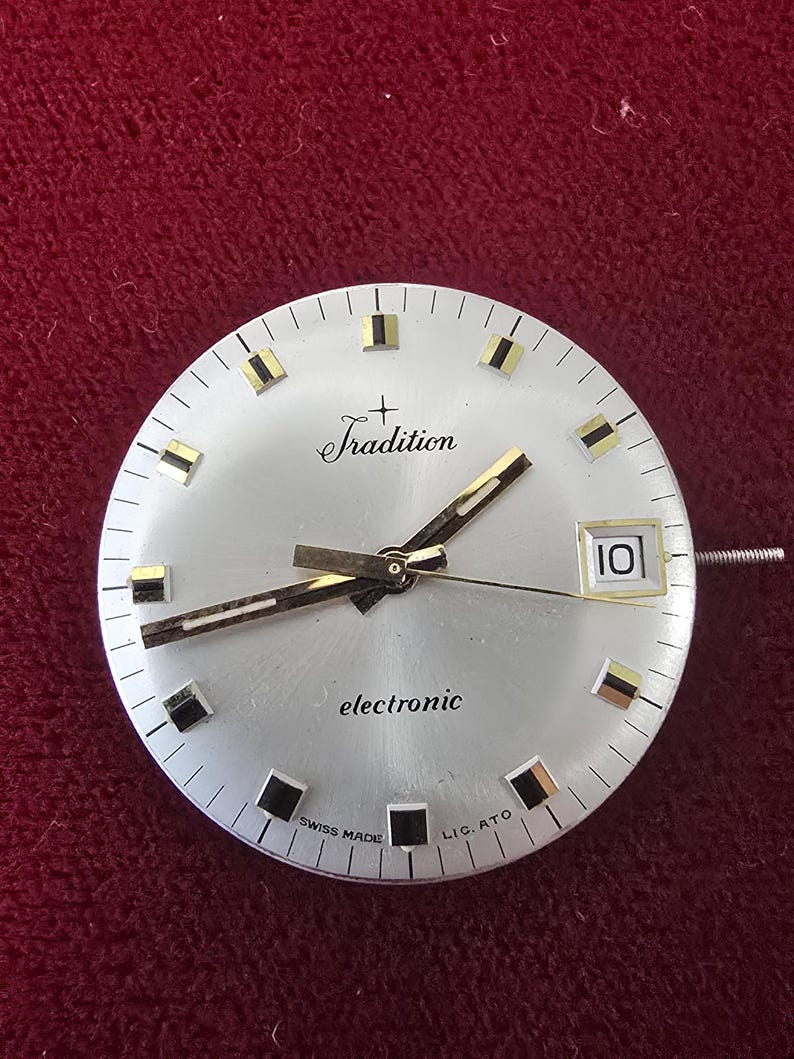 Puede incluir: Esfera de reloj plateada con las palabras "Tradition" y "electronic" impresas. La esfera del reloj tiene manecillas y marcadores dorados, con una ventana de fecha que muestra el n&uacute;mero "10". Las palabras "SWISS MADE" tambi&eacute;n est&aacute;n impresas.