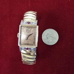 May include: A vintage Bulova watch with a rectangular face and a gold-tone band. The watch face is a light champagne color with black hands and numerals. The band has a decorative design with red accents. A quarter is next to the watch.