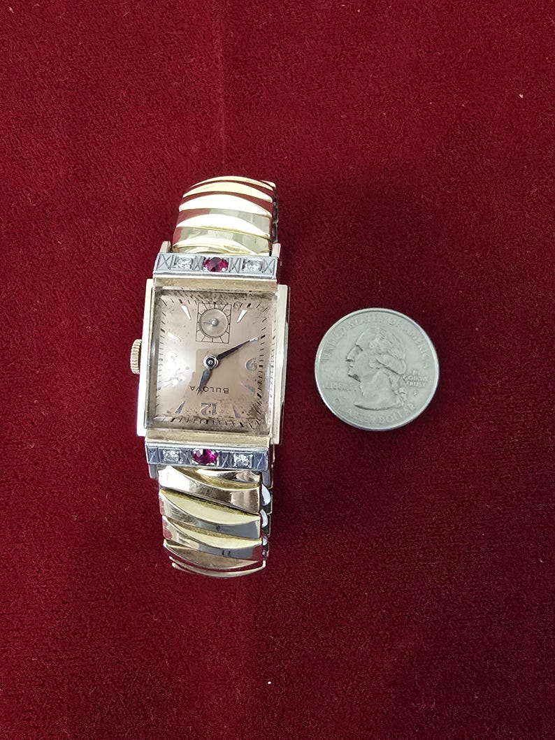 May include: A vintage Bulova watch with a rectangular face and a gold-tone band. The watch face is a light champagne color with black hands and numerals. The band has a decorative design with red accents. A quarter is next to the watch.