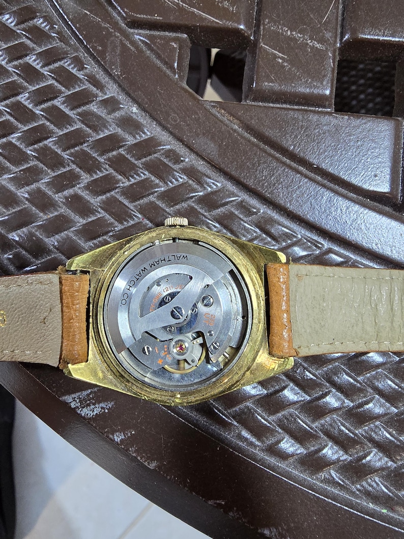 May include: A vintage gold-toned Waltham watch with a tan leather band. The watch's back is visible, showcasing the intricate mechanical movement and the inscription "WALTHAM WATCH CO."
