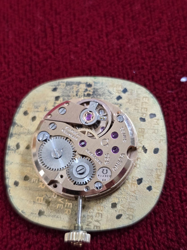 Pu&ograve; includere: Primo piano di un movimento di orologio Omega vintage. Il meccanismo dorato presenta ingranaggi intricati, gioielli e il logo Omega. Il movimento dell'orologio &egrave; montato su una base dorata con testo sui bordi. Lo sfondo &egrave; un tessuto rosso scuro.