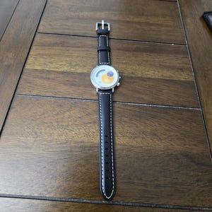 Op de afbeelding: Een polshorloge met een zwarte leren band met witte contraststiksels en een zilveren gesp. De ronde wijzerplaat heeft een witte achtergrond met een gele cirkelvormige afbeelding en een kleine blauwe cirkel, gevat in een zilverkleurige metalen kast.
