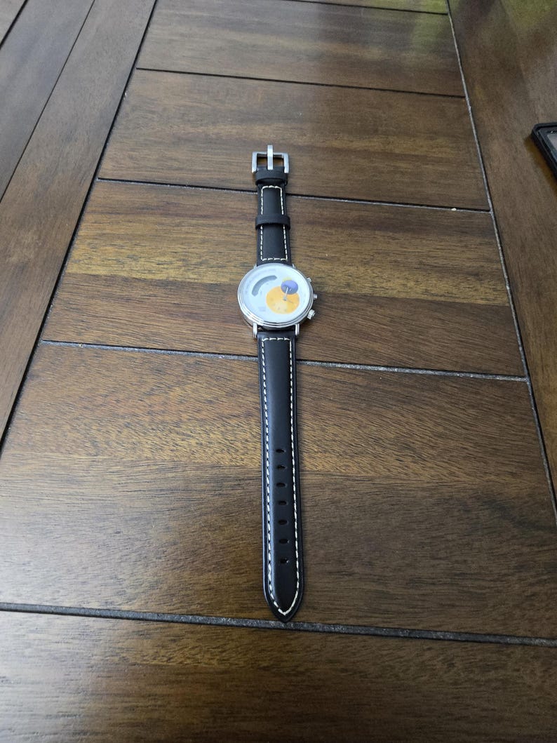 Op de afbeelding: Een polshorloge met een zwarte leren band met witte contraststiksels en een zilveren gesp. De ronde wijzerplaat heeft een witte achtergrond met een gele cirkelvormige afbeelding en een kleine blauwe cirkel, gevat in een zilverkleurige metalen kast.