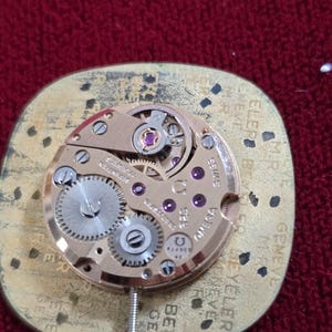 Pu&ograve; includere: Primo piano del movimento di un orologio Omega vintage, che mostra ingranaggi intricati, gioielli e il logo Omega. Il meccanismo dorato &egrave; posto su una base dorata con testo. Un esempio di ingegneria di precisione.