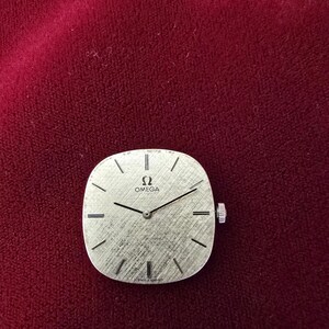 Pu&ograve; includere: Un quadrante dell'orologio Omega argentato con un quadrante strutturato e lancette nere. L'orologio ha una forma quadrata con angoli arrotondati e una piccola corona sul lato destro. Il quadrante presenta il logo Omega e il testo Swiss Made.