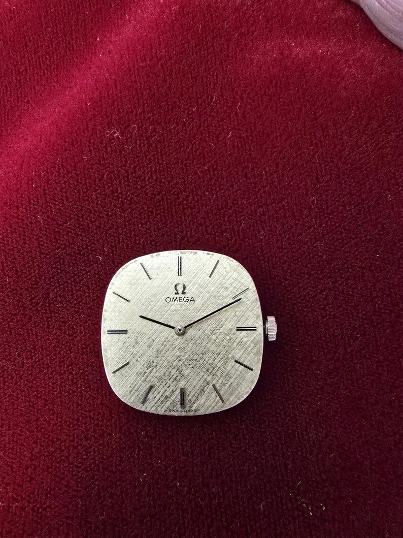 Pu&ograve; includere: Un quadrante dell'orologio Omega argentato con un quadrante strutturato e lancette nere. L'orologio ha una forma quadrata con angoli arrotondati e una piccola corona sul lato destro. Il quadrante presenta il logo Omega e il testo Swiss Made.