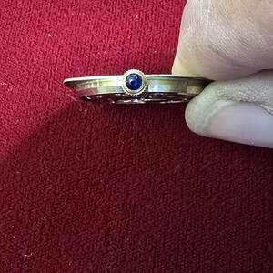 May include: A gold-toned watch component with a small, round, dark blue stone set in the center. The watch part is held by a gloved hand against a red background.