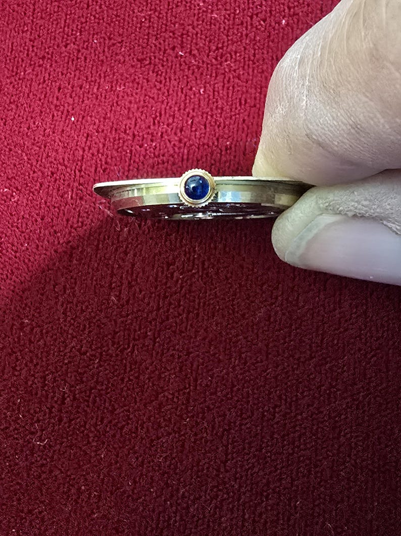 May include: A gold-toned watch component with a small, round, dark blue stone set in the center. The watch part is held by a gloved hand against a red background.
