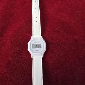 May include: A white Casio digital watch with a white leather strap. The watch face displays the time. The watch is on a red background.
