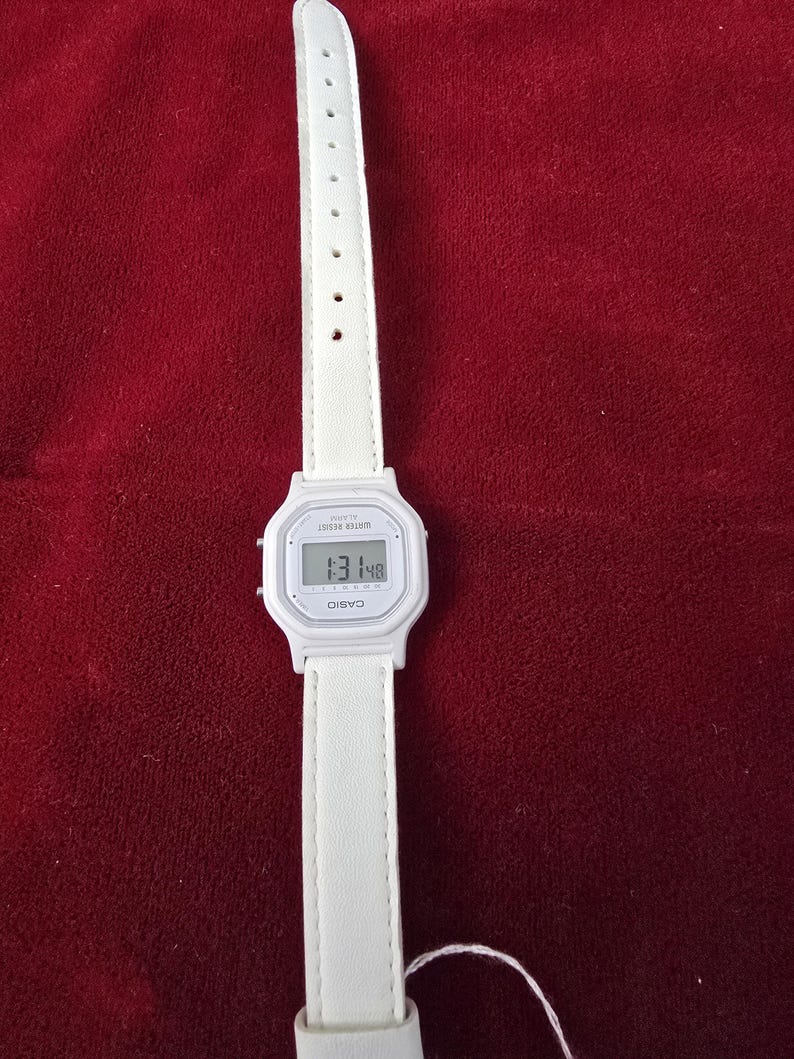May include: A white Casio digital watch with a white leather strap. The watch face displays the time. The watch is on a red background.