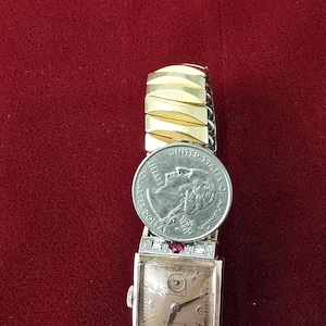 May include: Vintage Bulova watch with a rectangular face, gold-tone band, and a quarter coin placed on top. The watch face has a light-colored background with black hands and numerals. Red gemstones are set on the watch.